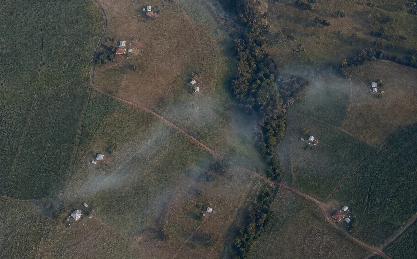 Vista aérea de zona rural ao amanhecer: lavouras, estradas de terra, várias sedes de propriedade espalhadas pelo vale e fumaça se espalhando sobre a mata que acompanha o curso d'água.